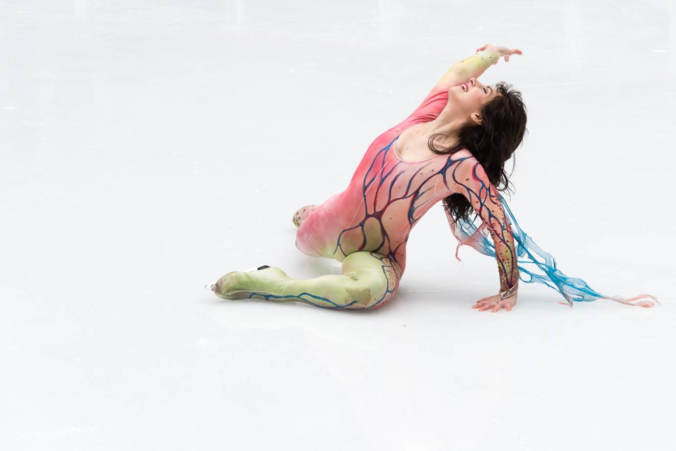 An ice dancer in a pastel colored unitard, lays on the ice and passionately raises her arm to the sky